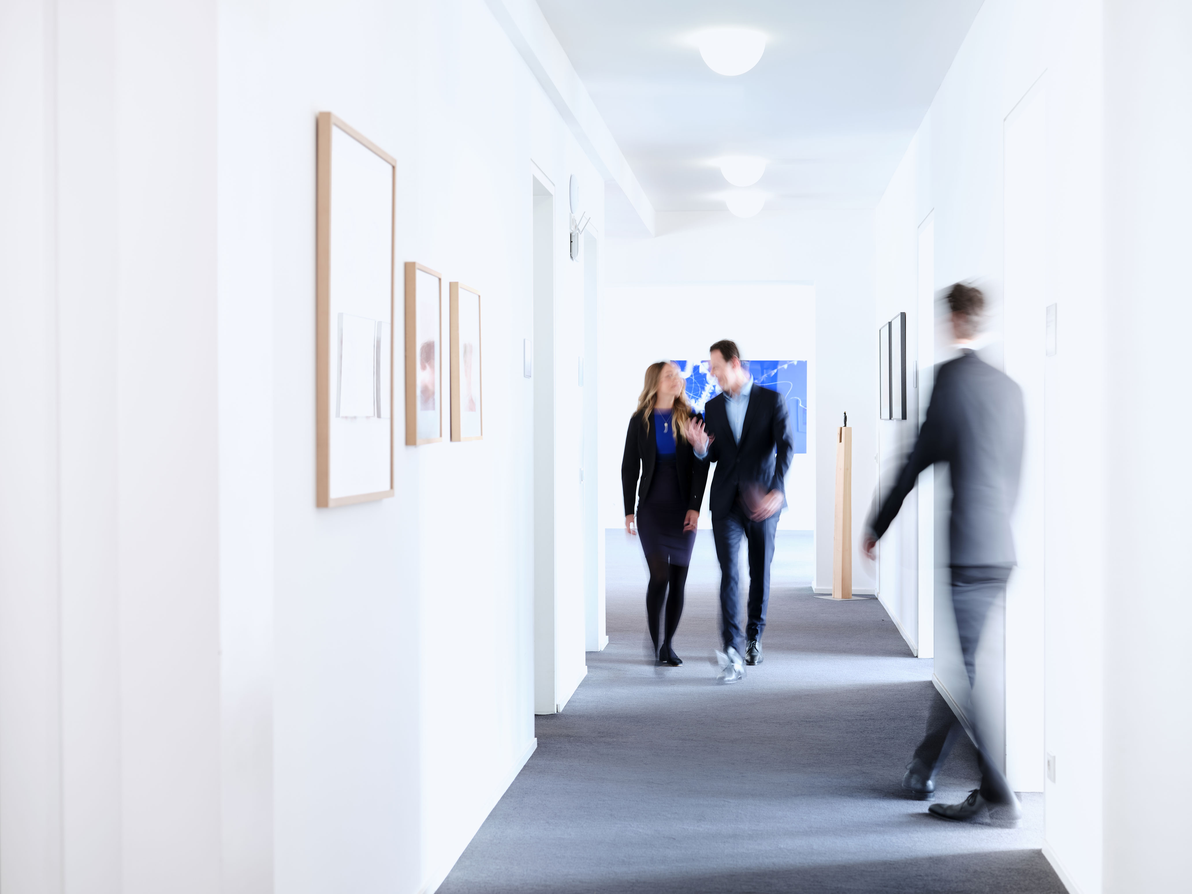 Auf dem Imagebild ist ein Flur im Kölner Büro zu sehen. Rechts und Links sind die Türen zu den einzelnen Büros. Auf dem Bild sieht man zwei Personen, welche zur Kamera laufen und sich dabei unterhalten. Eine weitere Person geht rechts aus einem Büro raus zum Ende des Flurs.