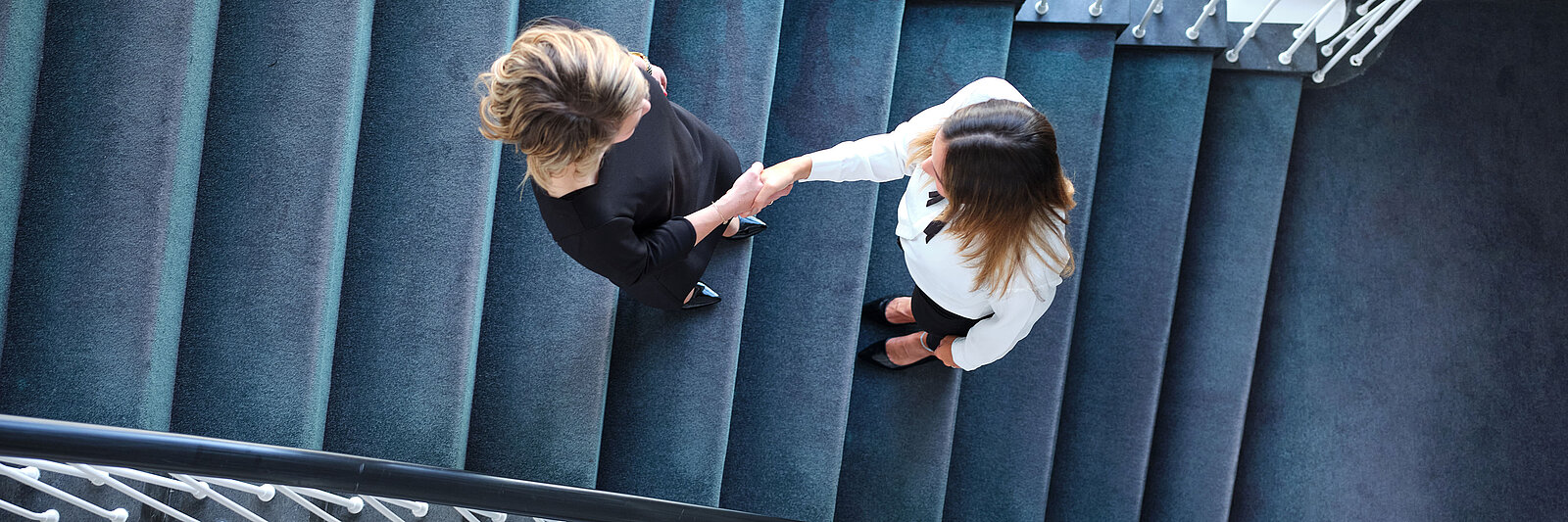 Auf dem Imagebild wurde das Treppenhaus des Kölner Standorts von oben fotografiert. In der Mitte der Treppen stehen zwei Personen, welche sich mit einem Händeschütteln begrüßen.